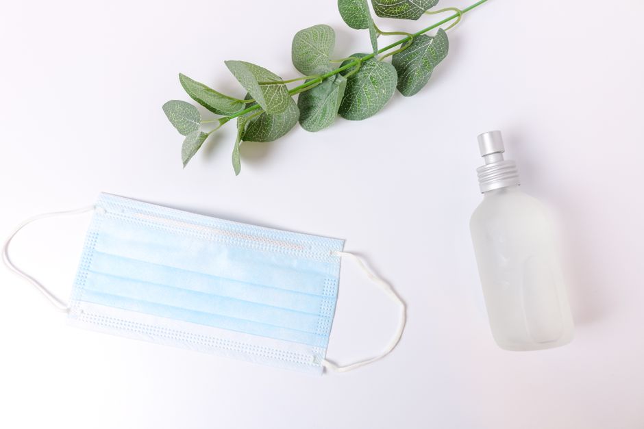 A flat lay of a face mask, sanitizer spray bottle, and green leaves on a white surface.